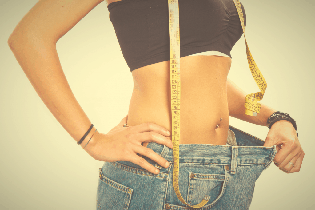 Fitness After 50: How To Lose Belly Fat Woman with tape measure around her neck holding waist of oversized jeans to show belly fat loss.